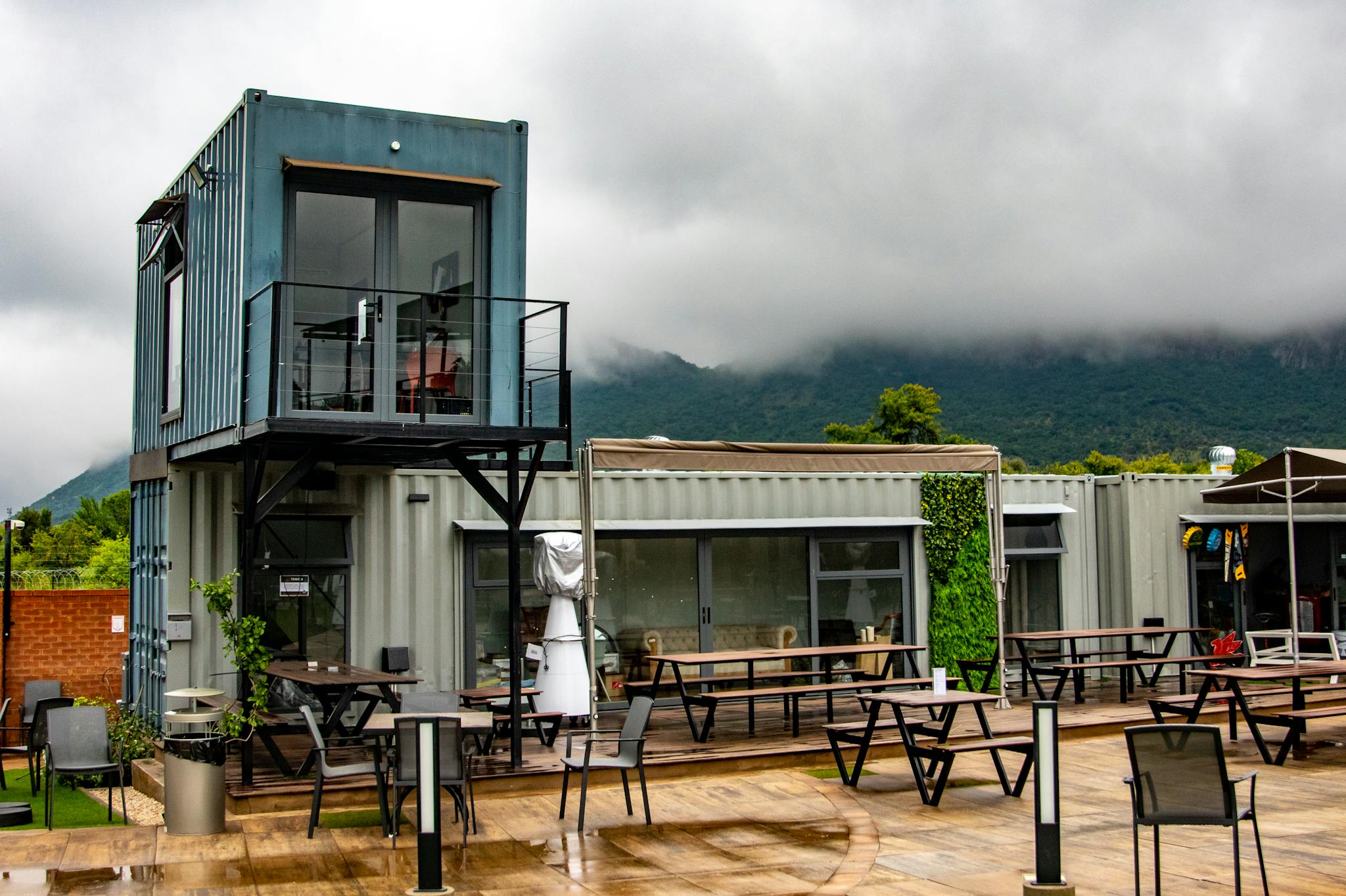 A contemporary container cafe set amidst lush greenery and cloudy mountains, offering a unique outdoor experience.